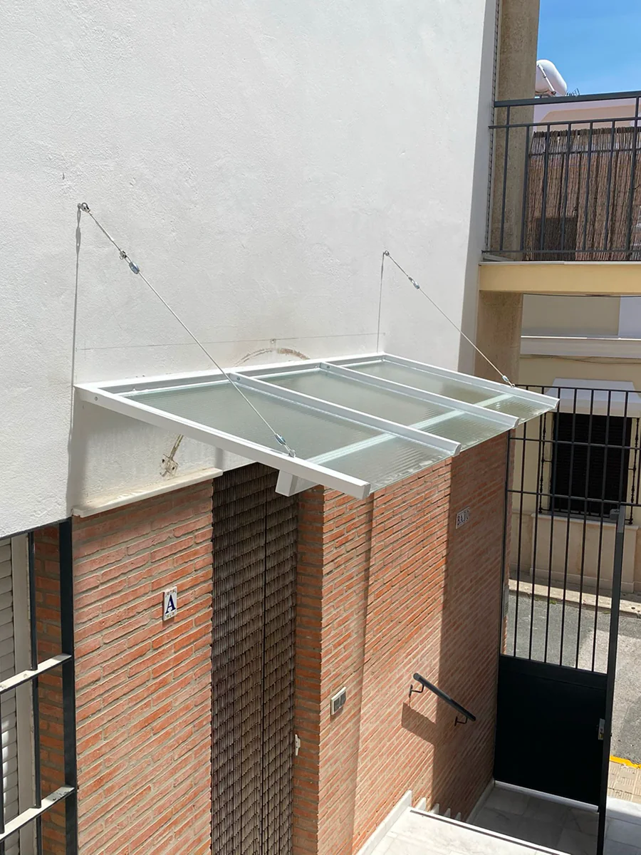 Marquesina volada de aluminio blanco y vidrio laminado sobre acceso en Sevilla